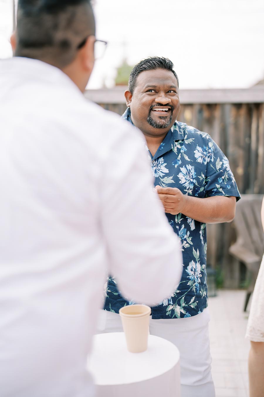 Smiling man in a floral shirt engaged in conversation at an outdoor gathering.