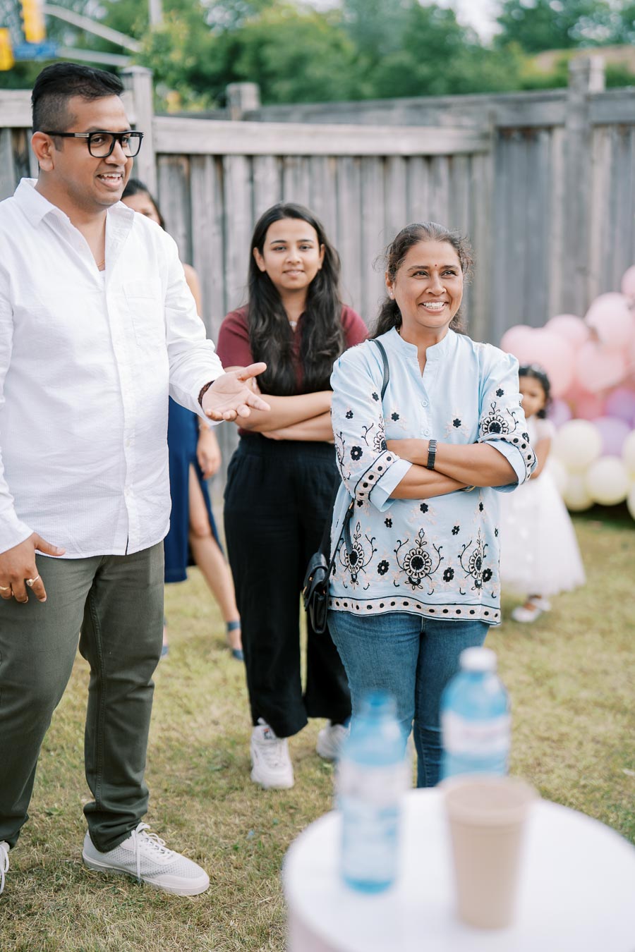 A group of people enjoying an outdoor event, with two women and one man in casual attire smiling and engaging in conversation, set against a wooden fence and balloons in the background.