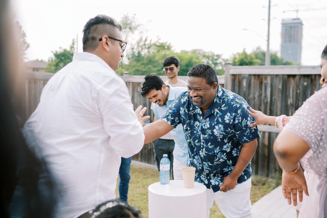 Group of friends enjoying an outdoor gathering, laughing and playing a game around a small white table in a backyard setting.