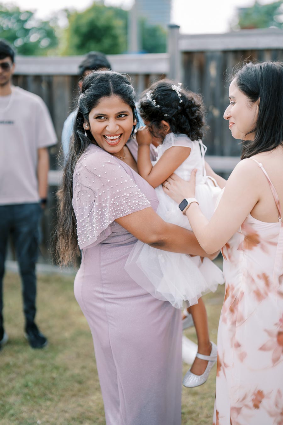 A joyful woman in a lavender dress holds a young child outdoors, smiling brightly, while another woman looks on in a floral dress, with a wooden fence and greenery in the background.