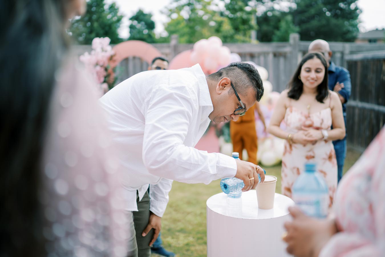 Man in a white shirt pouring water into a cup at an outdoor gathering with blurred guests and colorful decorations in the background.