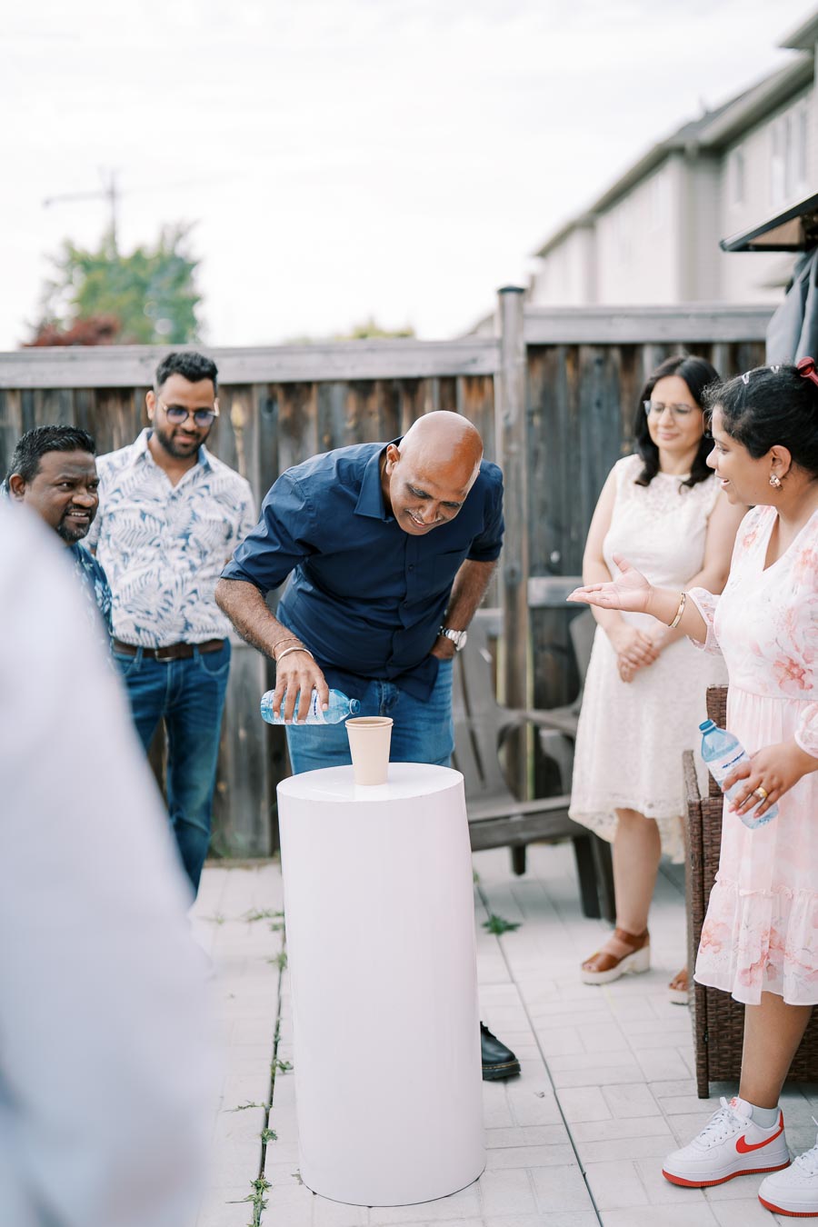 A group of people gathered outdoors around a cylindrical table, engaging in a fun challenge with a cup and water bottle.