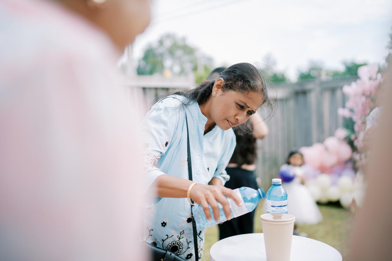 Woman pouring water into a cup outside during a garden party, surrounded by balloons and soft, colorful decor.