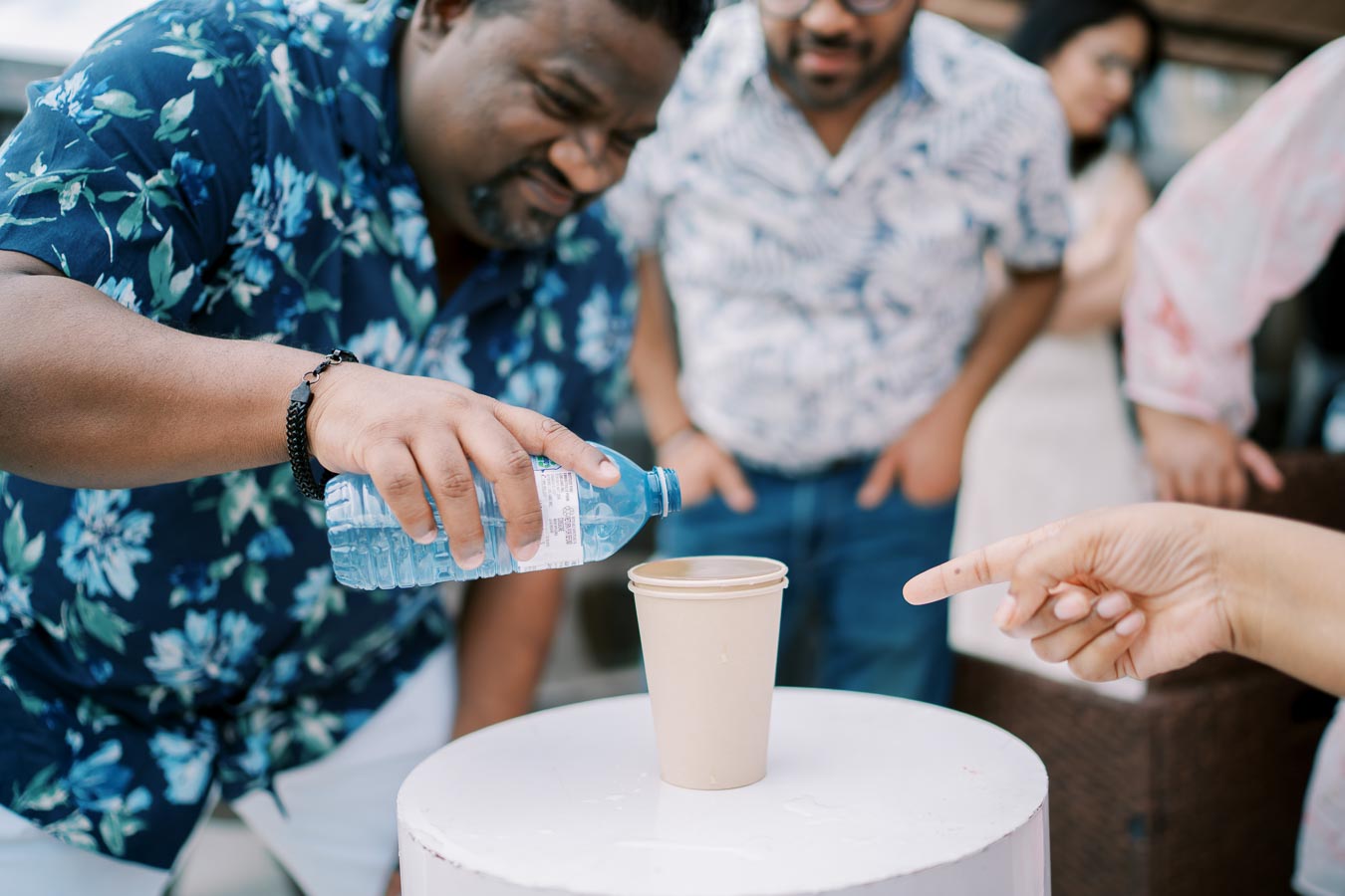 Man in a floral shirt pours water from a bottle into a cardboard cup, with people watching in the background during an outdoor gathering.
