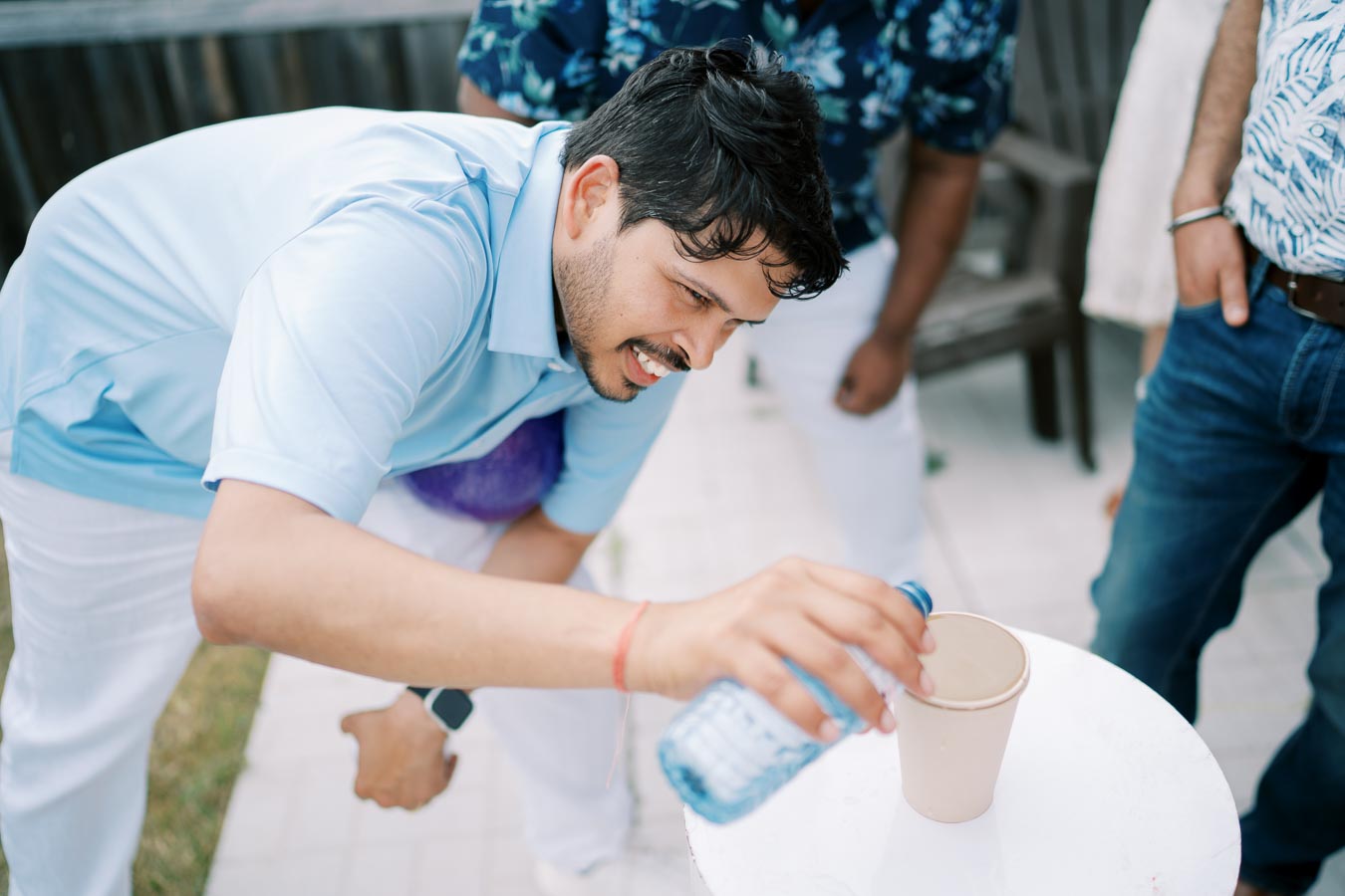 A smiling man in a light blue shirt pours water from a bottle into a paper cup at an outdoor gathering, surrounded by casually dressed people in a relaxed setting.