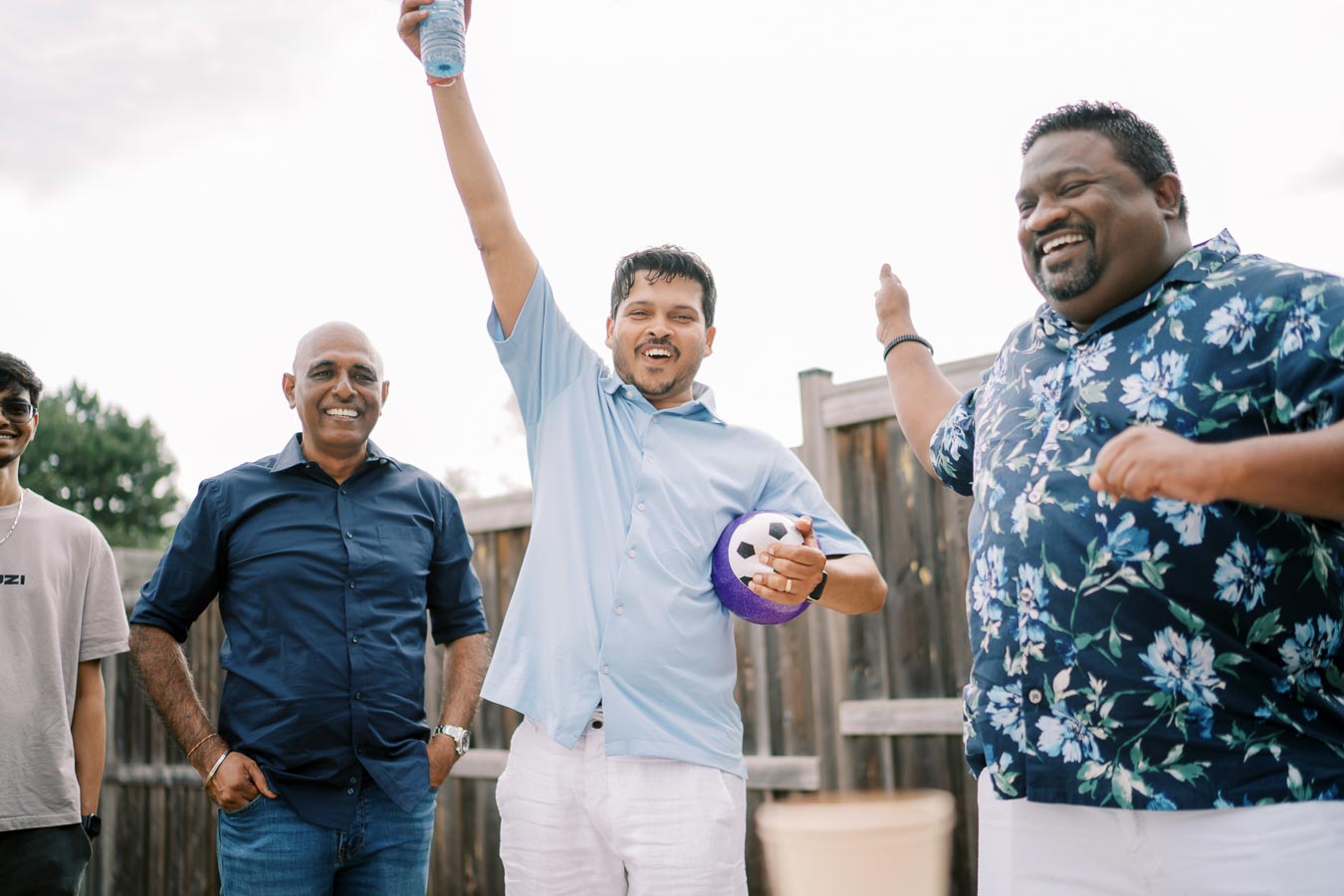 A group of four men having fun outdoors, with one man raising a drink and holding a small soccer ball, smiling and enjoying a casual gathering in a backyard setting, with a wooden fence and trees in the background.