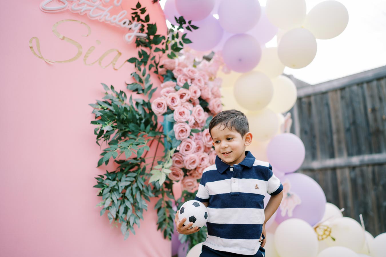 A young boy holding a mini soccer ball stands in front of a pink birthday backdrop decorated with pink roses and green leaves, surrounded by multicolored balloons.