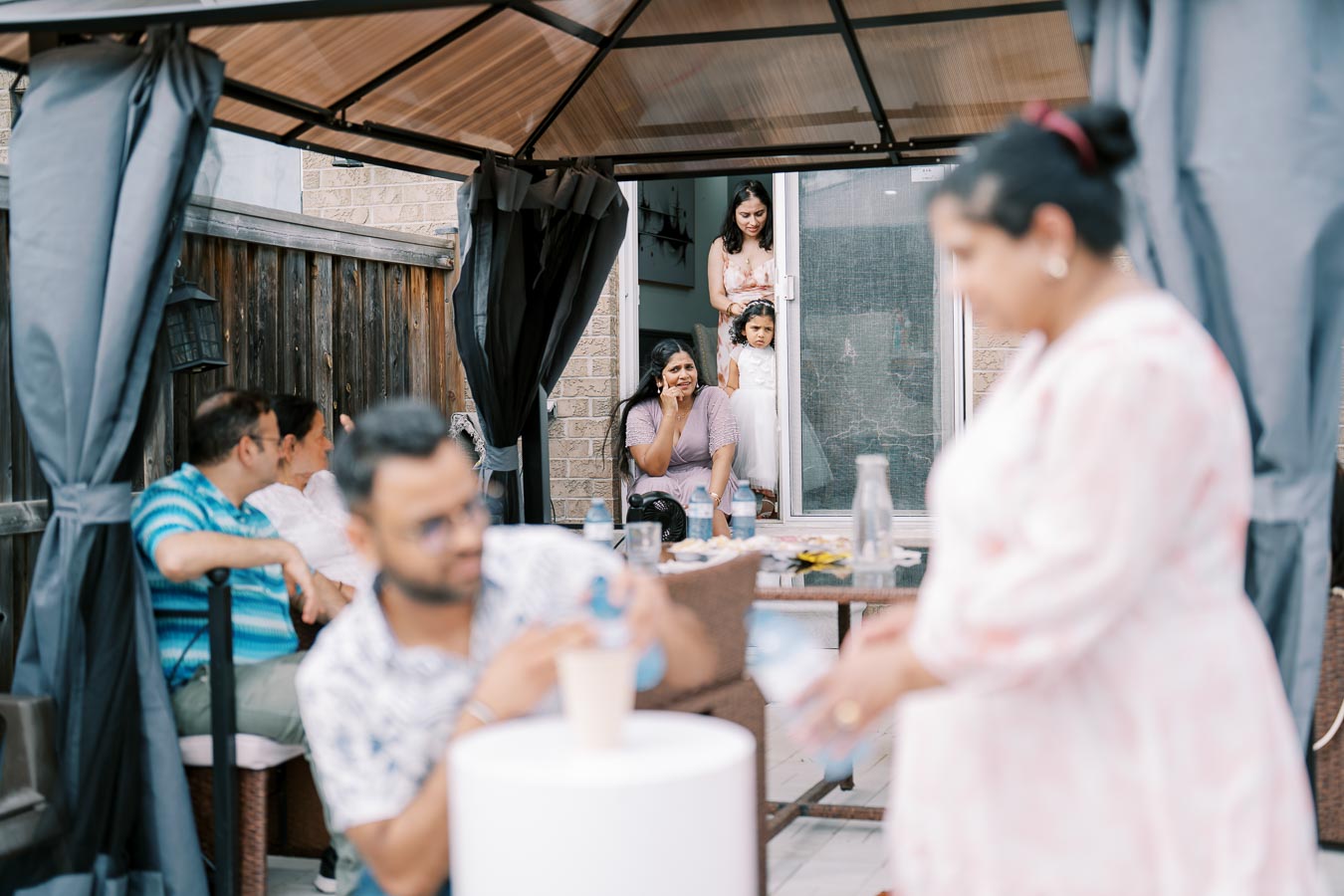 A group of people enjoying a backyard gathering under a canopy, with two women and a child at a sliding door, creating a relaxed and family-friendly atmosphere.