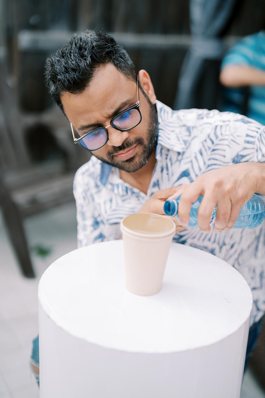 Man wearing blue glasses and patterned shirt pours water from a blue bottle into a paper cup outdoors.
