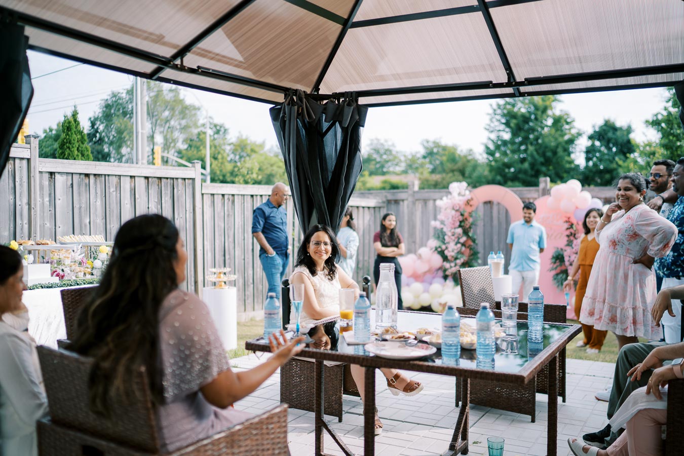 Group of people enjoying a backyard gathering under a gazebo, with a table filled with refreshments and a festive pink balloon backdrop.