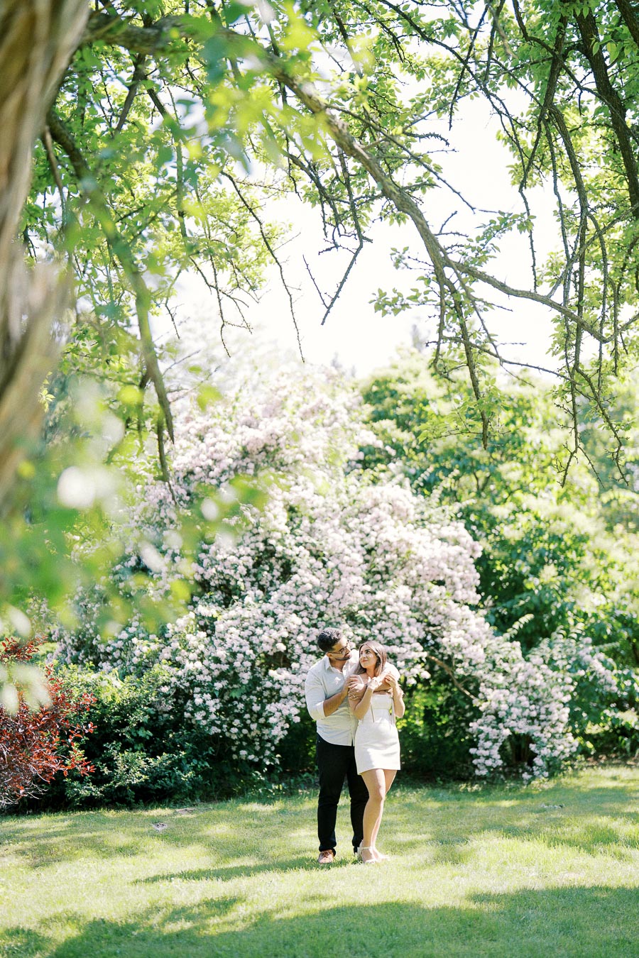 A romantic couple sharing an intimate moment in a sunlit garden, surrounded by lush greenery and blooming pink flowers.