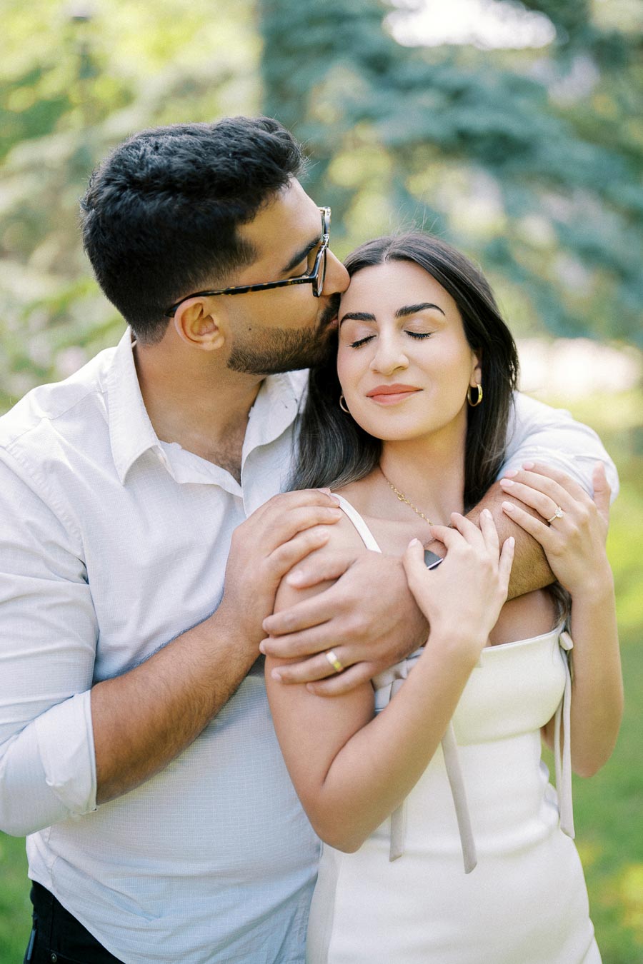 Romantic couple embracing in a lush green park, man wearing glasses and kissing woman on forehead, both smiling with eyes closed, conveying love and affection.