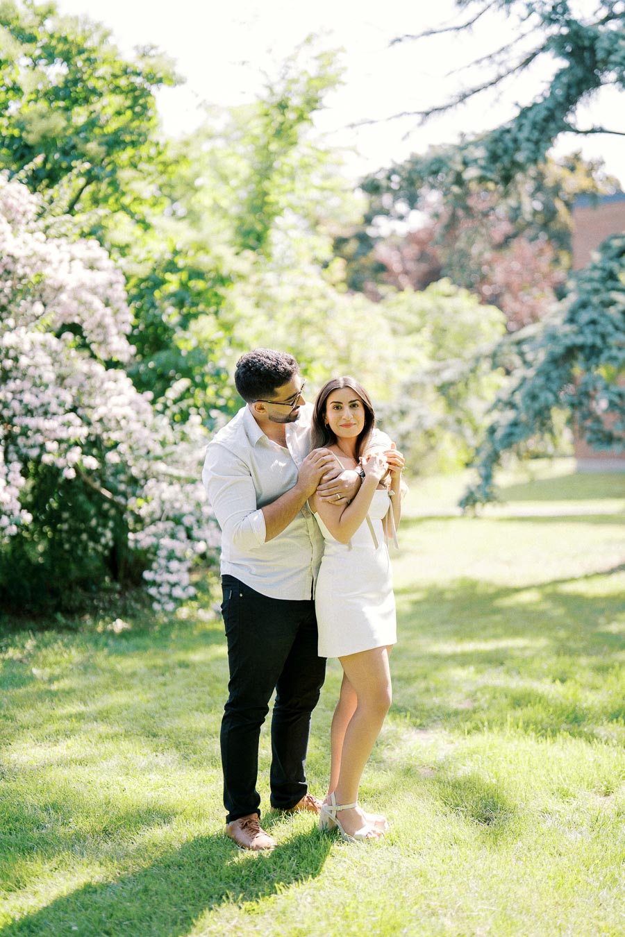 A couple embracing in a sunlit park, surrounded by lush greenery and blooming flowers, creating a romantic and serene atmosphere.