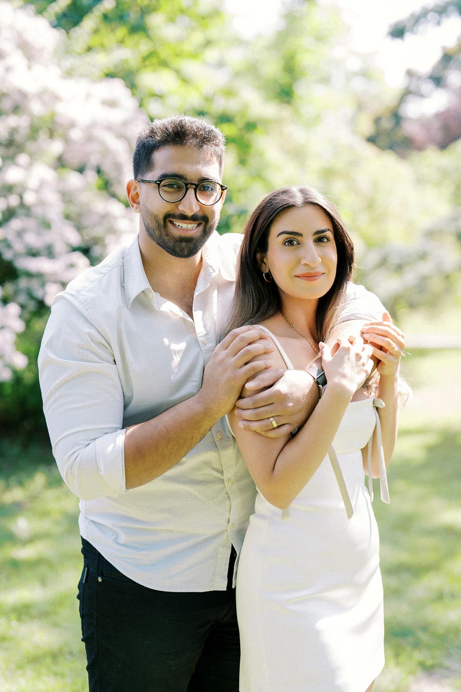 A couple embracing in a sunny garden with blooming flowers in the background, wearing casual yet elegant attire.
