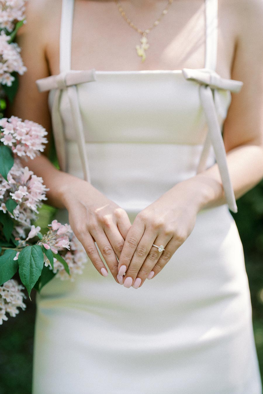 A woman in an elegant white dress stands near blooming pink flowers, highlighting a delicate gold necklace and a sparkling engagement ring on her left hand.