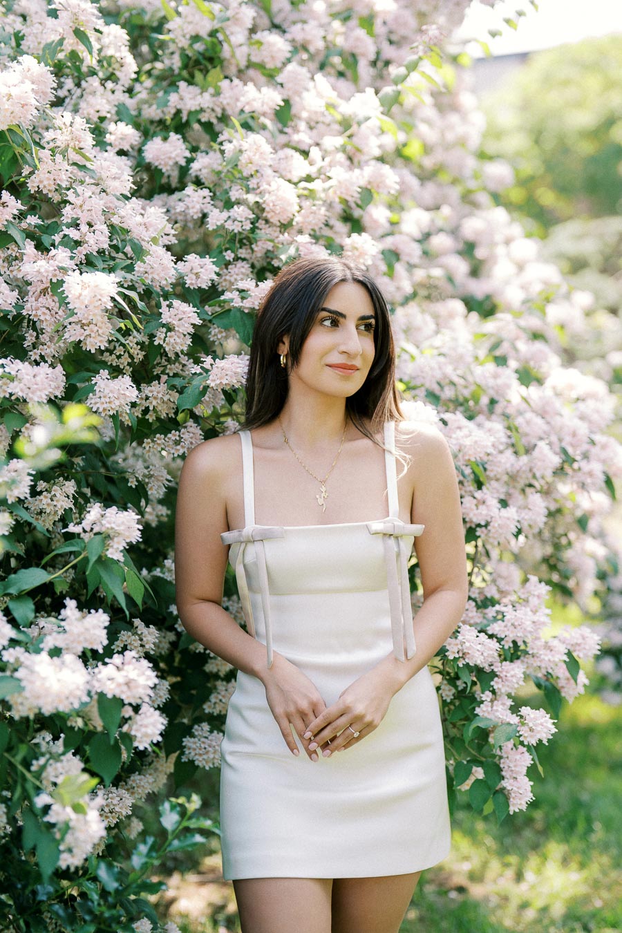 A woman in a white dress standing next to a blooming bush with light pink flowers in a garden setting.
