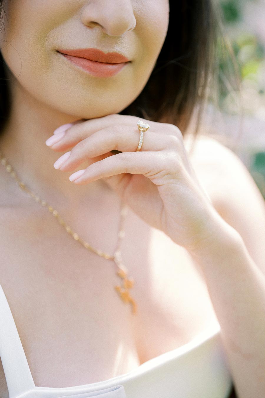 Close-up of a woman wearing a gold necklace and ring, with soft pink nail polish and a white top, in a sunlit outdoor setting.