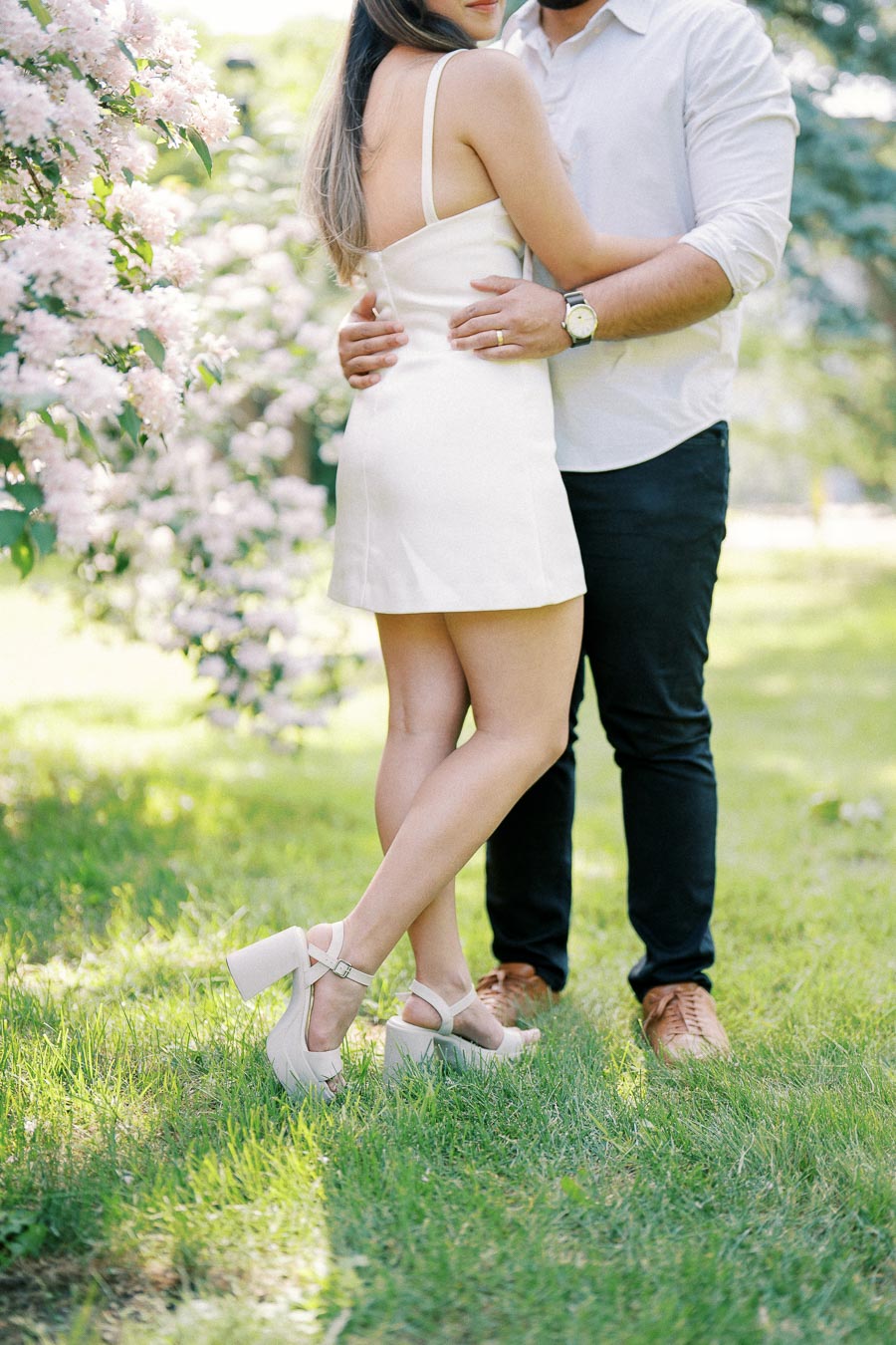 A couple embracing outdoors, woman in white dress and heels, surrounded by blooming flowers and greenery on a sunny day.