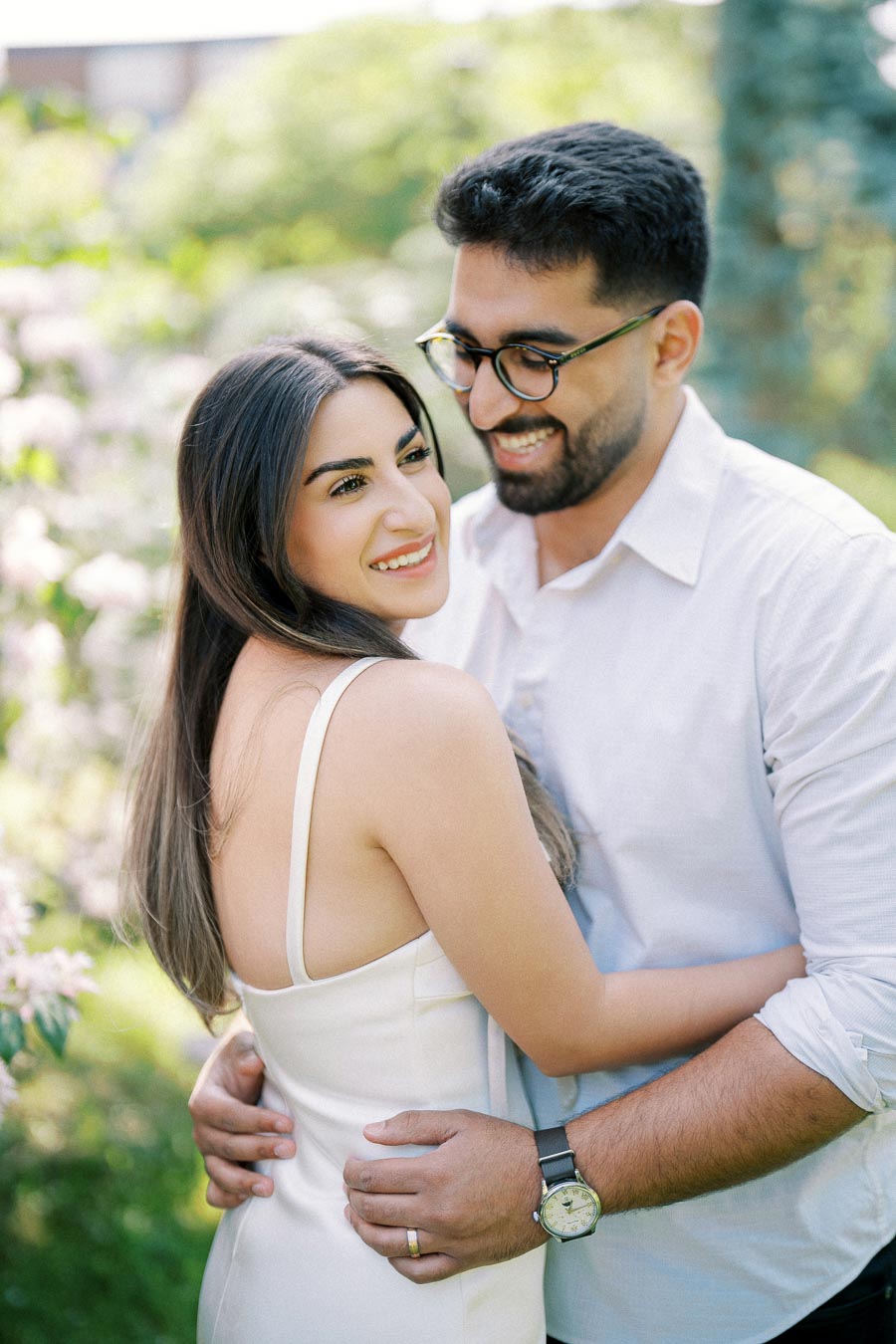 A happy couple embraces in a sunlit garden, with lush greenery and blooming flowers in the background, capturing a candid and romantic moment.