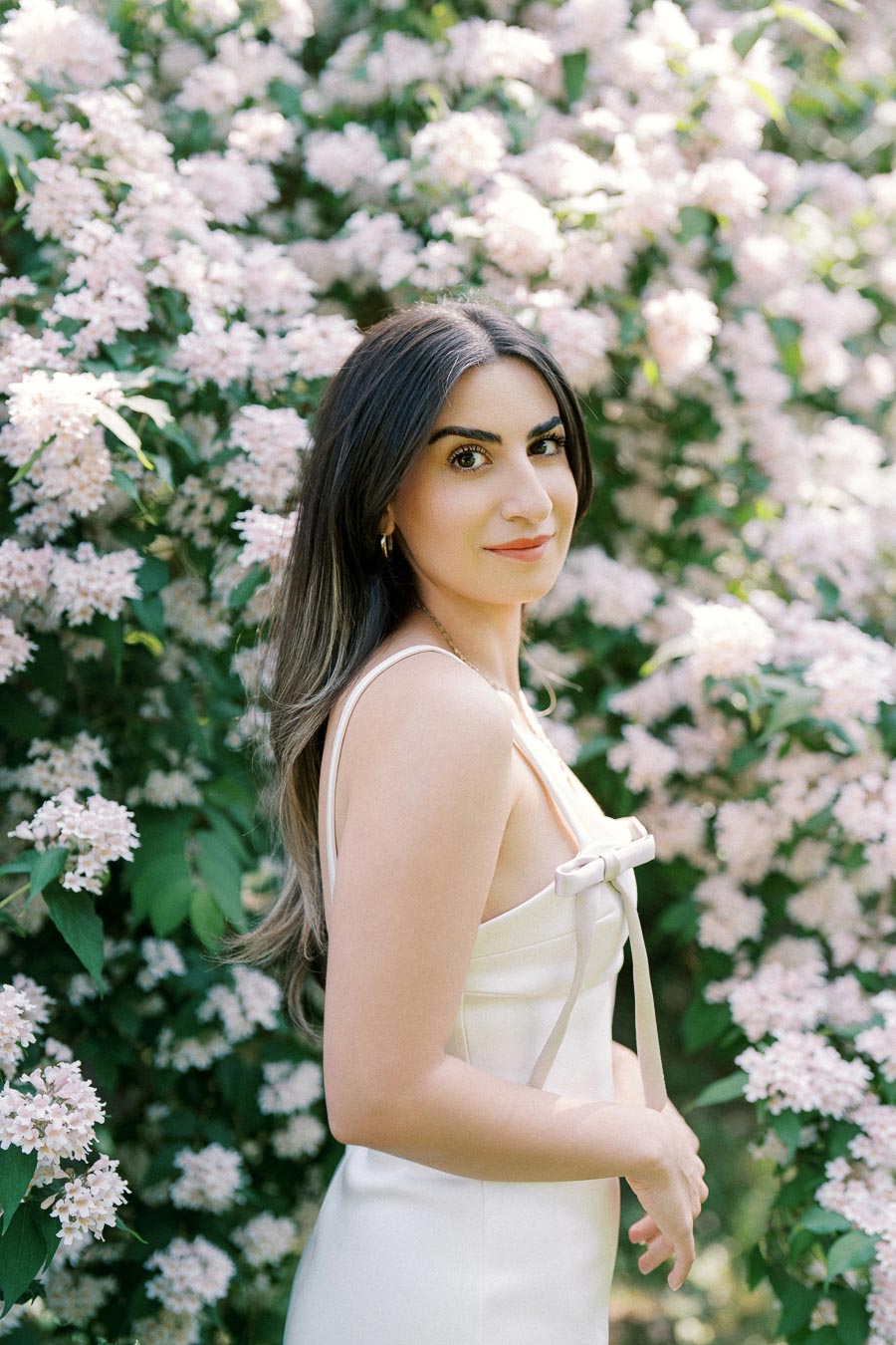 Woman in a white dress standing in front of a bush with blooming light pink flowers.