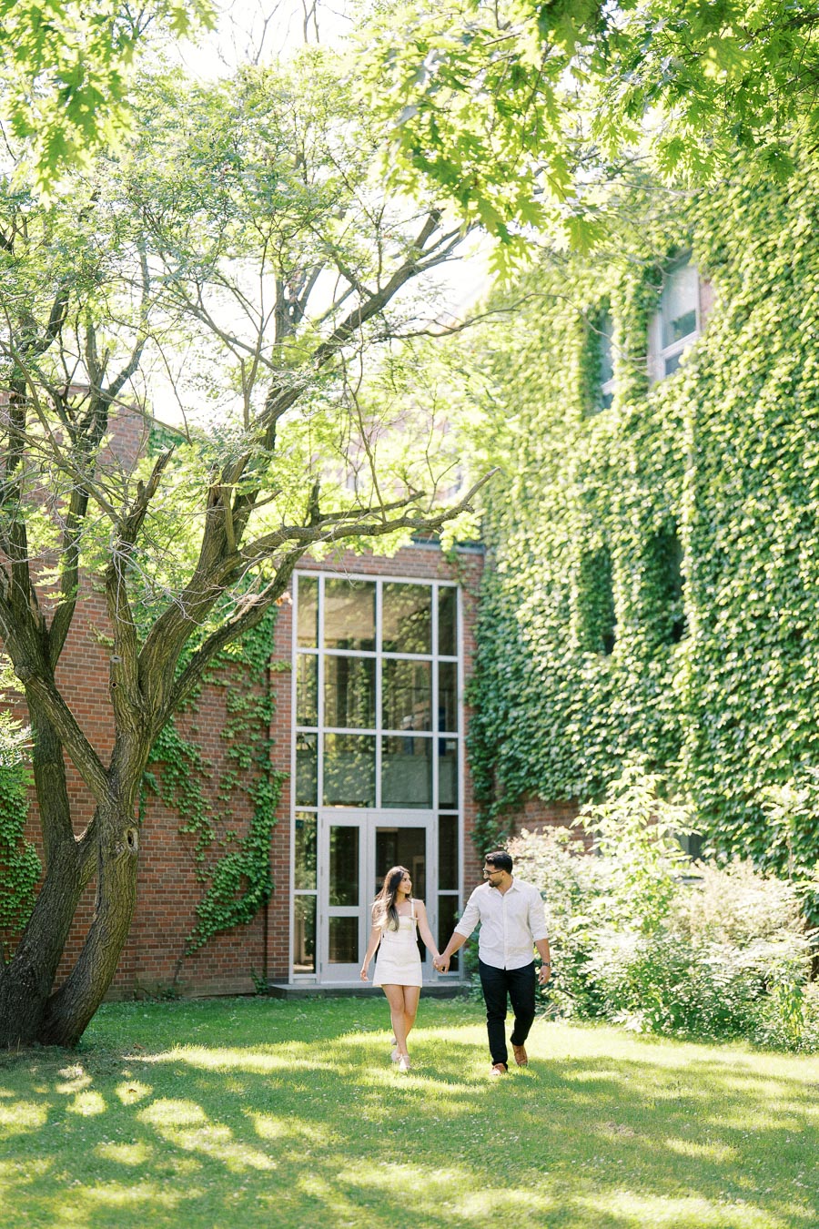 A couple holding hands and walking on a lush green lawn in front of a brick building covered with ivy, under a sunny, tree-filled sky.