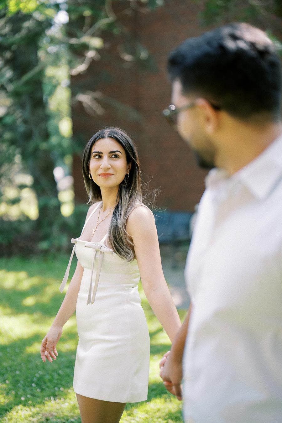 A couple holding hands outdoors, with a woman in a white dress looking at a man wearing a light-colored shirt. They are in a grassy garden setting with trees in the background.