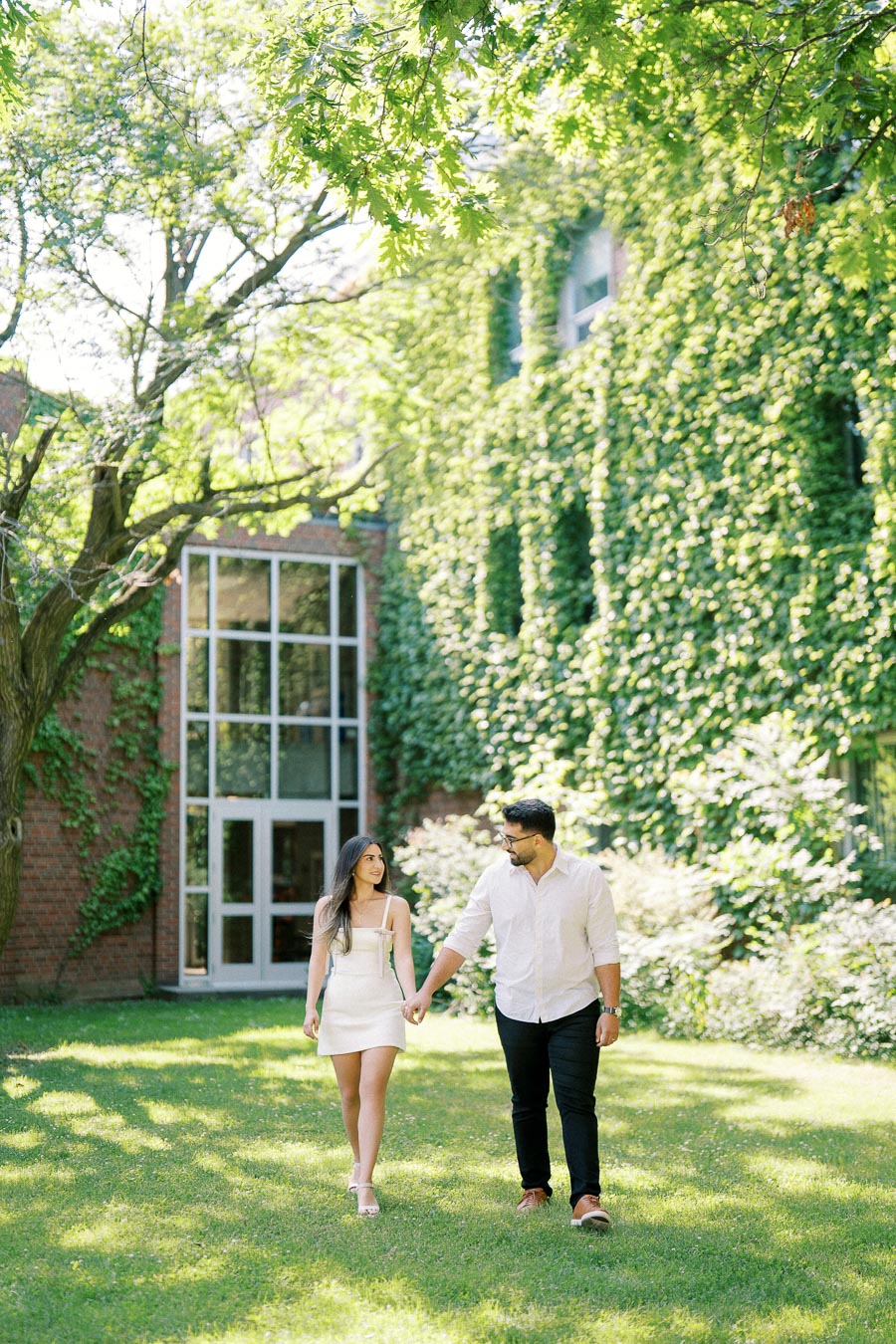 A couple holding hands and walking on a sunlit lawn in front of a building covered in green ivy, with trees providing shade.