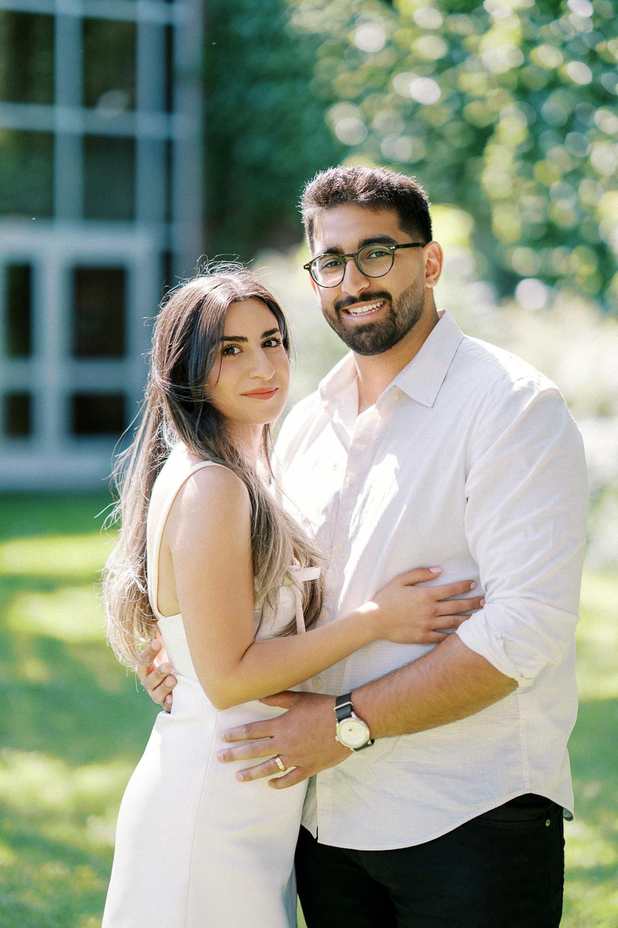 A couple embracing outdoors in a garden setting, with lush greenery and a building in the background. The man is wearing a white shirt and glasses, while the woman is in a white dress.