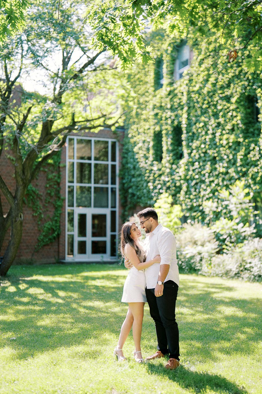 A couple embracing and smiling in a lush garden setting, surrounded by greenery and ivy-covered buildings. The sunlight filters through the trees, adding a romantic and peaceful ambiance to the scene.
