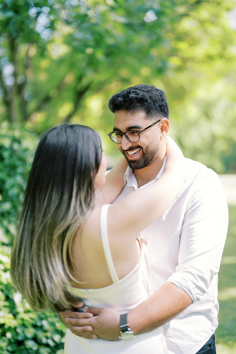 A couple embracing and smiling at each other in a sunlit, leafy park setting.
