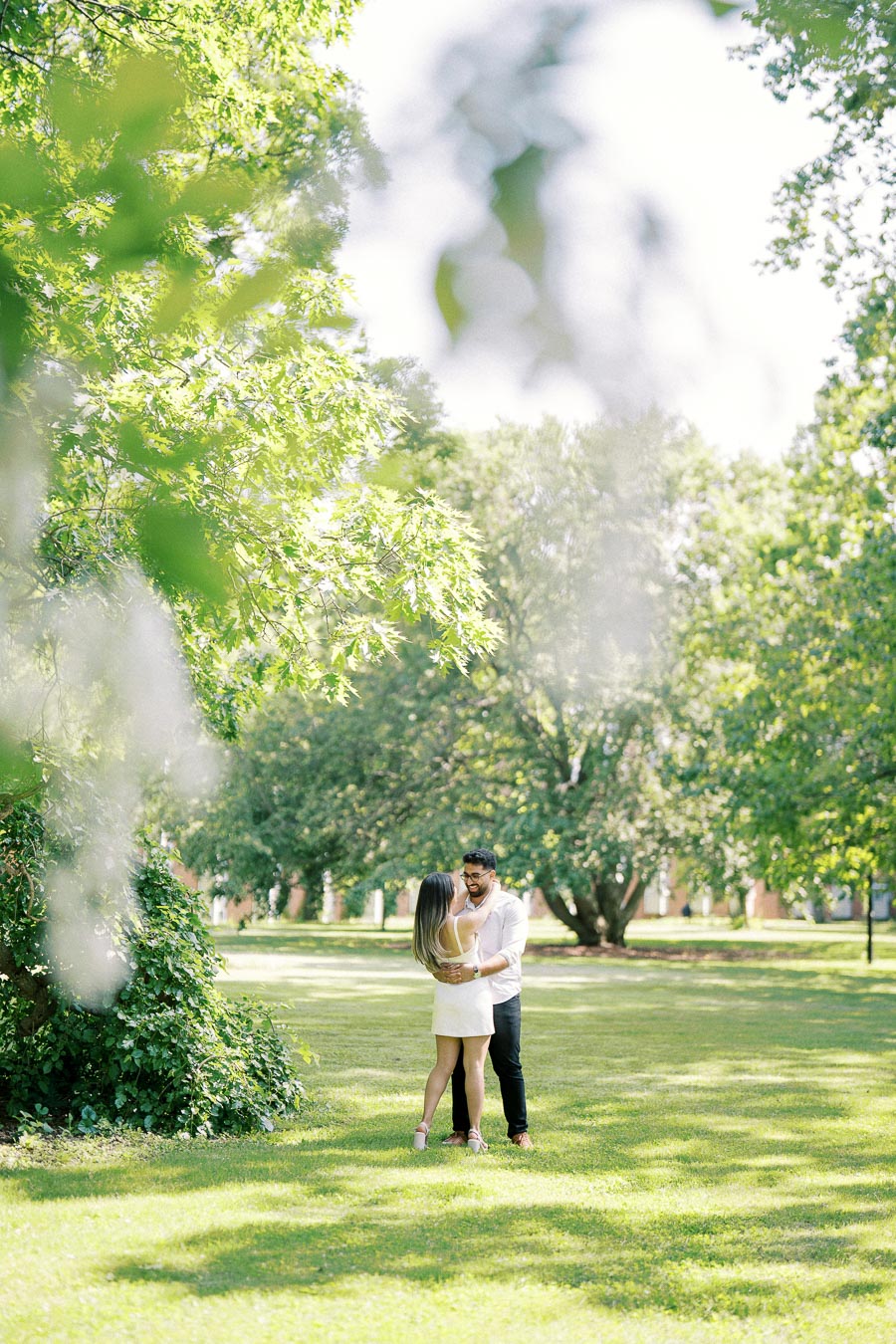 A couple embracing under a vibrant green tree in a sunny park.