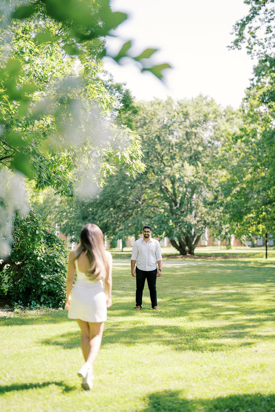 A woman in a white dress walks towards a man in a white shirt and black pants in a sunny, green park setting, surrounded by lush trees.