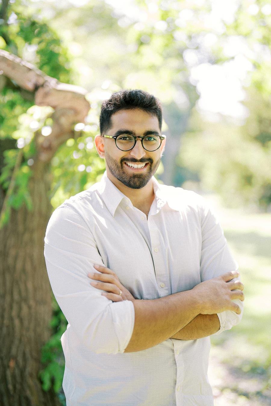 A smiling man with glasses and a beard, wearing a white shirt, stands with arms crossed in a sunny outdoor setting, surrounded by greenery and trees.