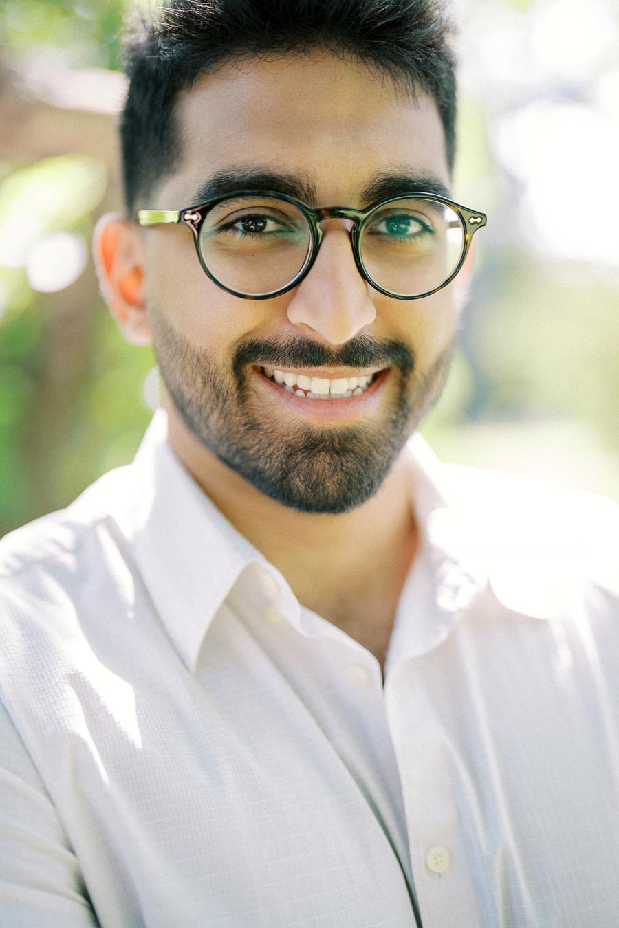 A smiling man with glasses and a beard wearing a white shirt, standing outdoors with sunlight and greenery in the background.