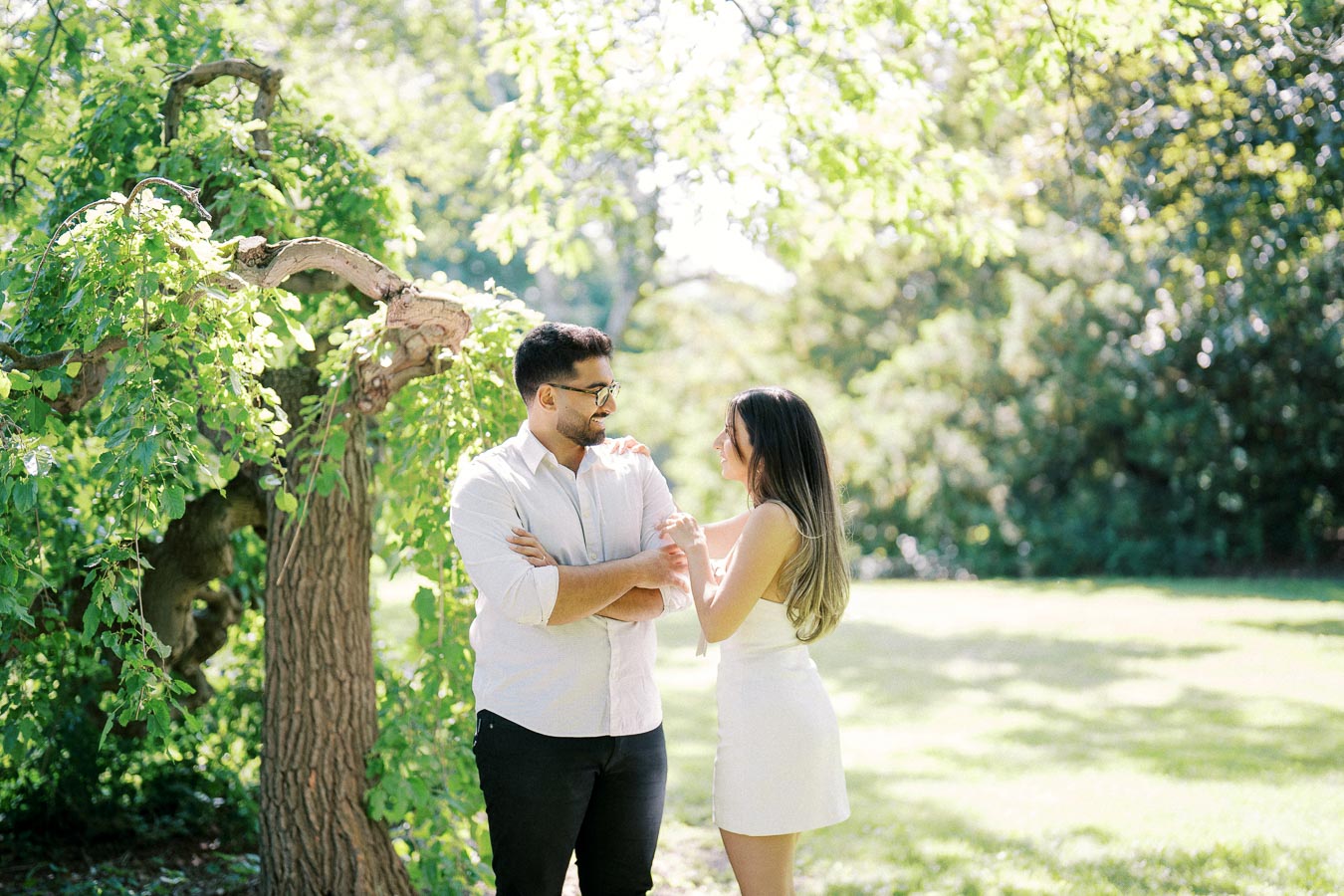 Couple standing in a sunlit park, smiling under a tree with lush green leaves.