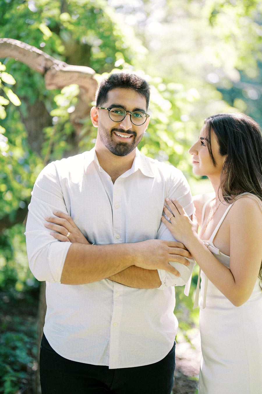 A couple standing in a sunlit forest, with the man wearing glasses and a white shirt, smiling while the woman gazes at him with her hand on his arm, surrounded by lush greenery.