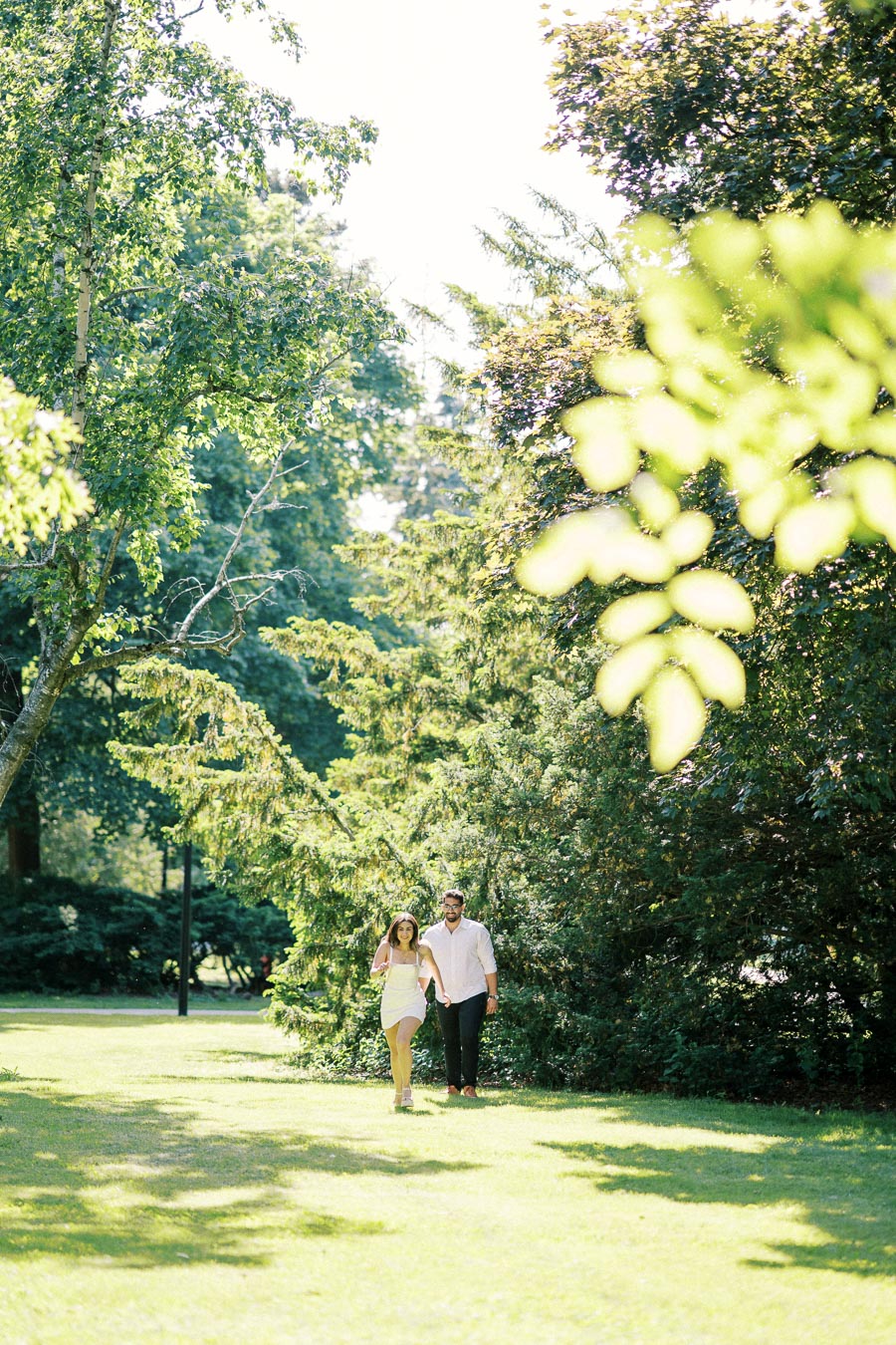 Couple walking through a sunlit, lush green park surrounded by tall trees and leafy branches.