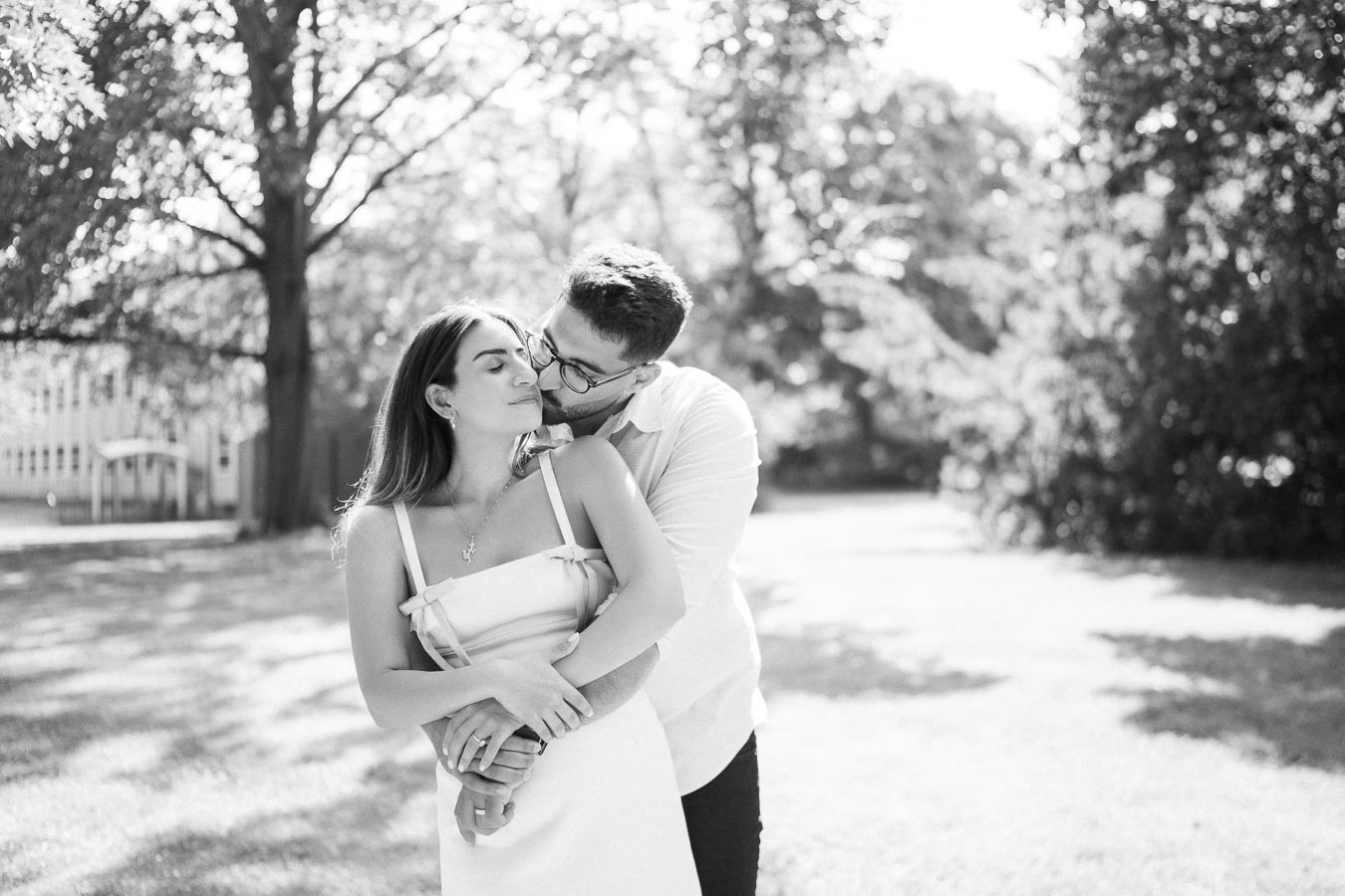 A romantic black and white image of a couple embracing in a sunlit park, surrounded by trees and greenery.