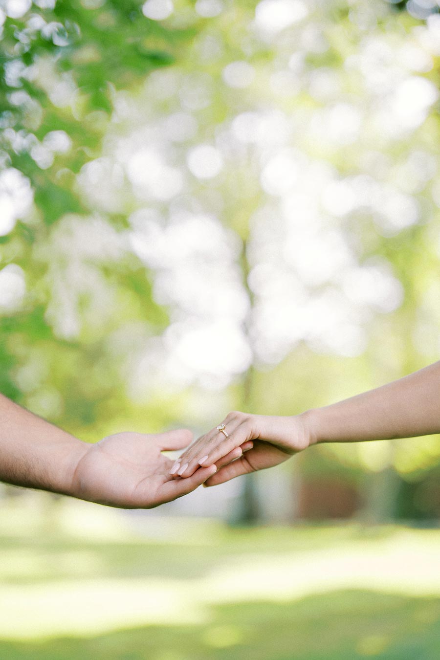 Two hands gently holding each other outdoors, with one wearing a gold engagement ring, set against a blurred green background.