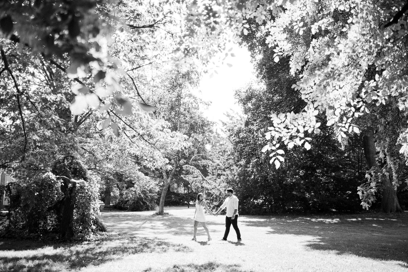 Black and white image of a couple walking hand in hand through a sunlit park, surrounded by lush trees and dappled sunlight.