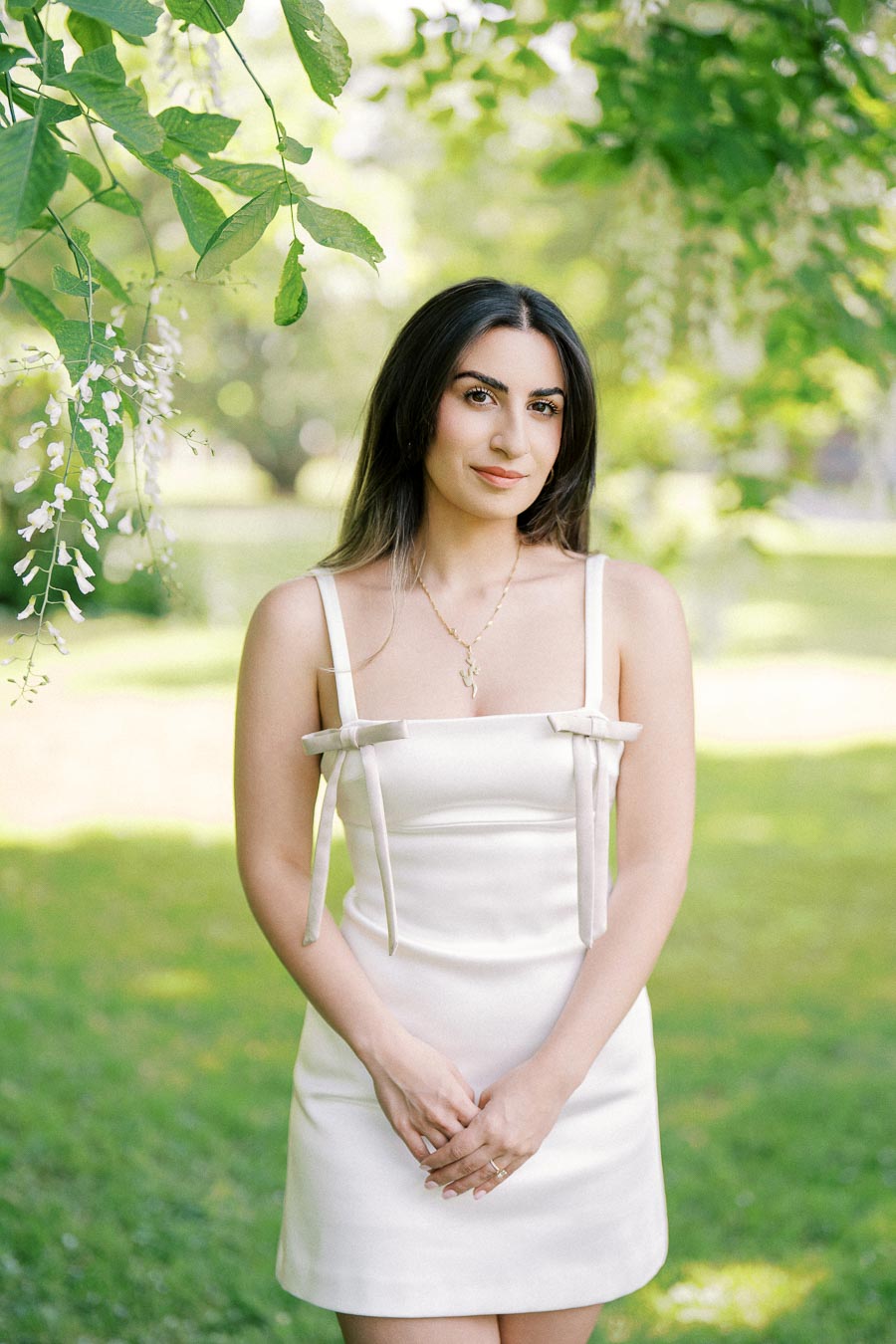 A woman in a white dress stands in a sunlit garden with green trees and hanging white flowers, looking at the camera.