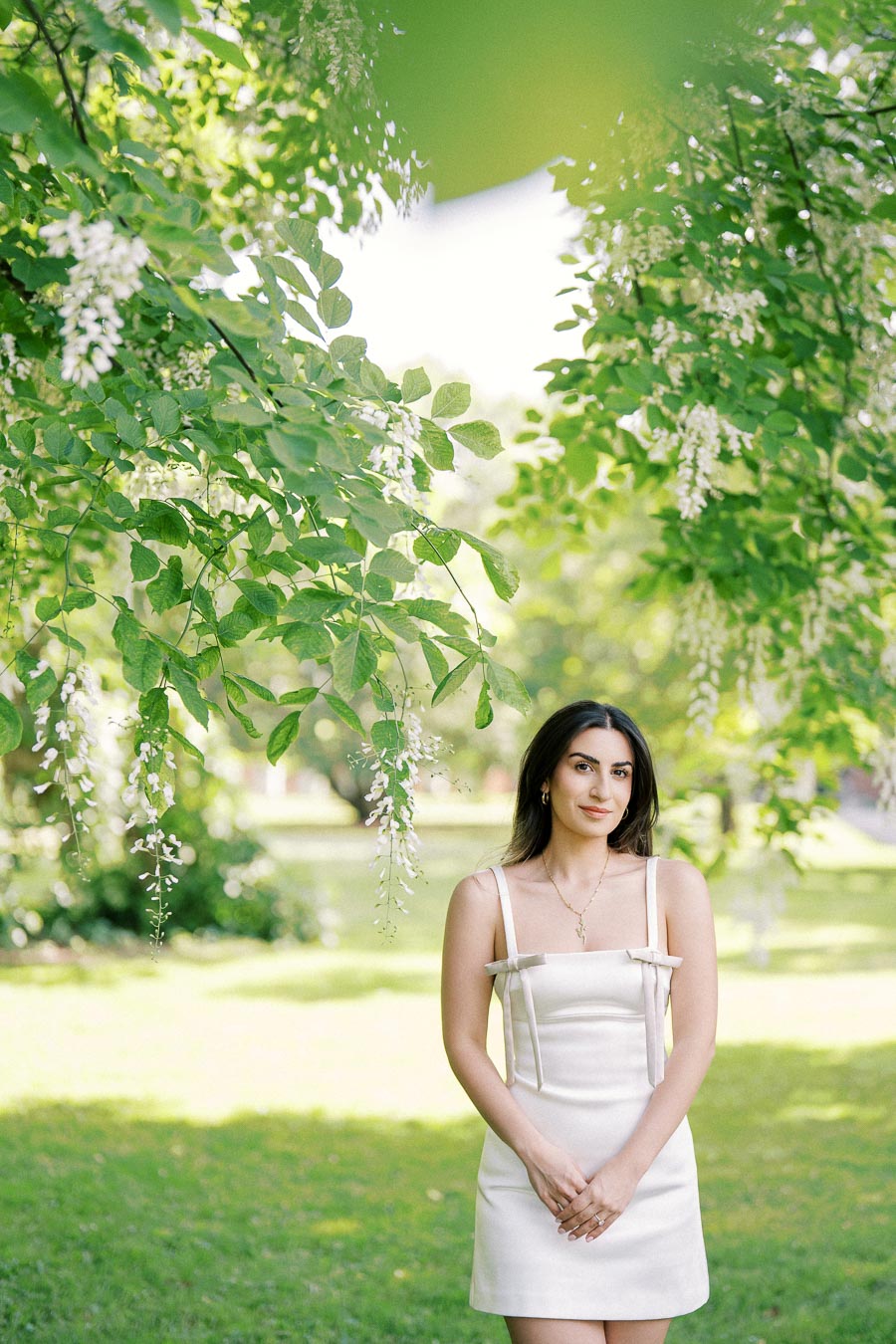 Woman in a white dress standing in a lush green park under hanging flowering branches, exuding a serene and elegant vibe.