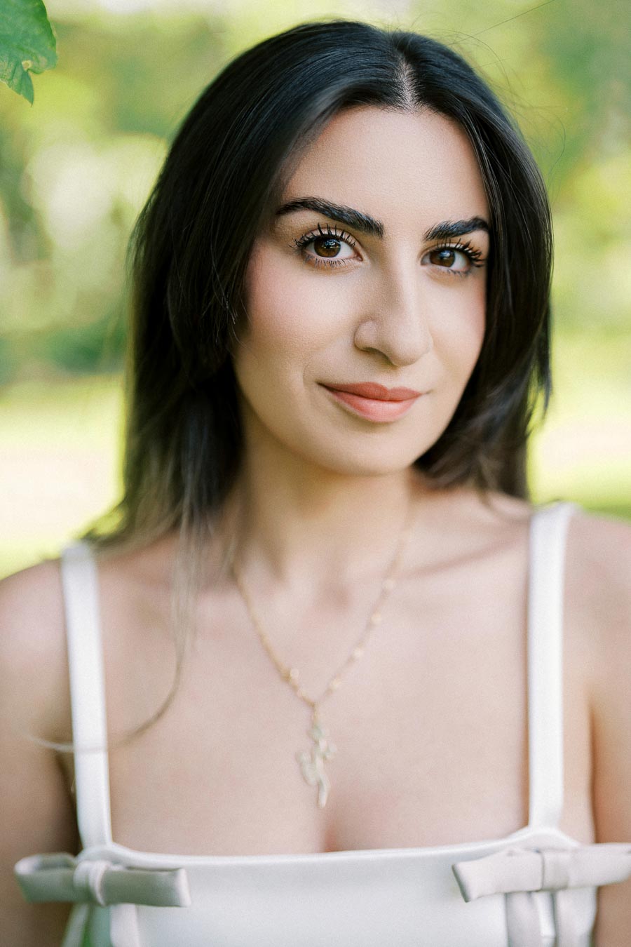 A woman with long dark hair wearing a white top and a necklace is smiling softly against a blurred green outdoor background.