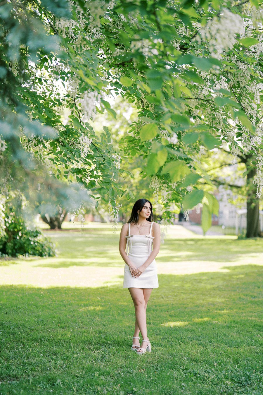 Young woman in a white dress standing on lush green grass under blossoming trees in a sunlit park.