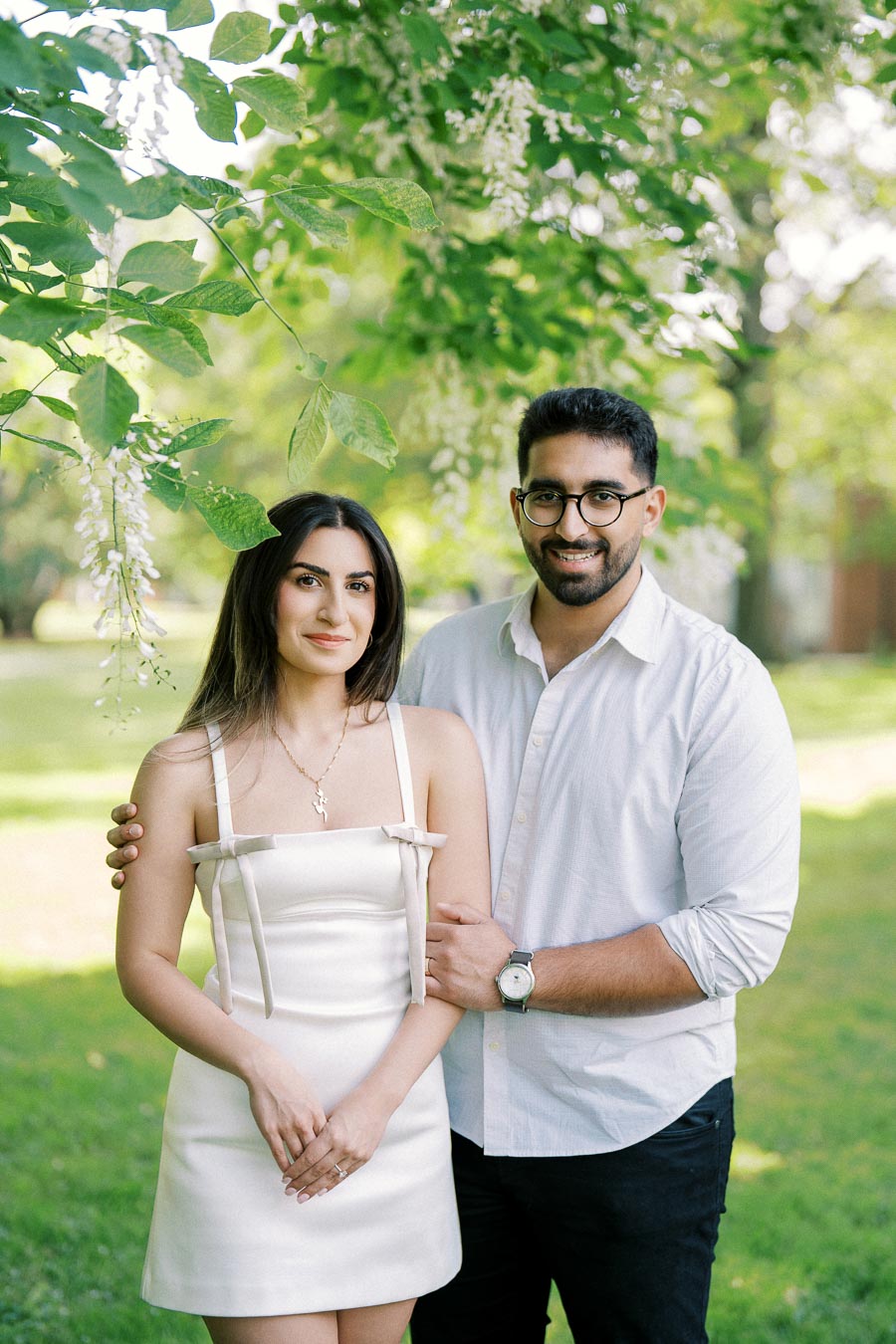A smiling couple standing together outdoors under lush green trees, dressed in light-colored clothing, enjoying a sunny day in the park.