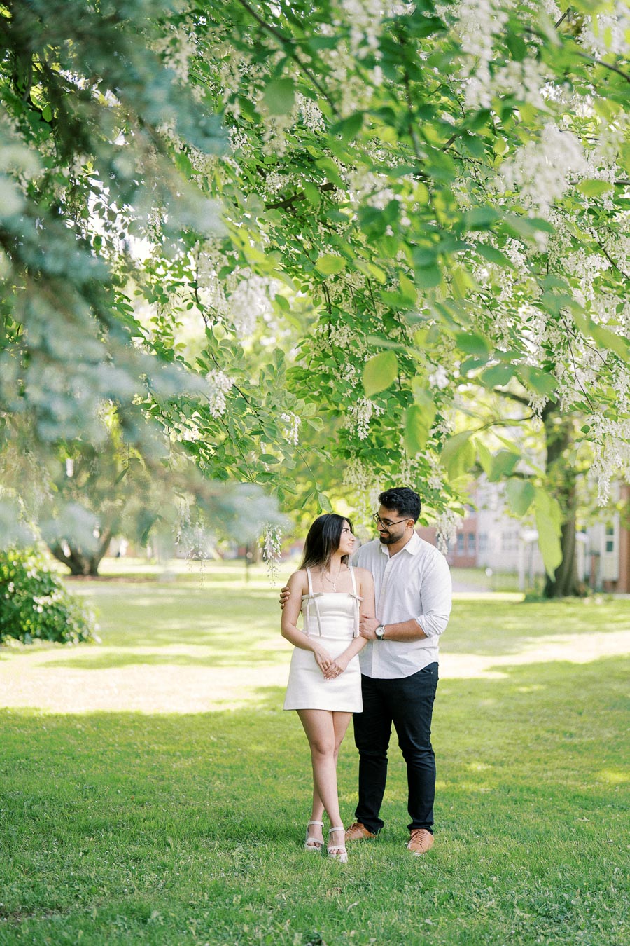 A couple embraces under lush green trees with white blossoms in a serene park setting, displaying affection and enjoying a sunny day outdoors.