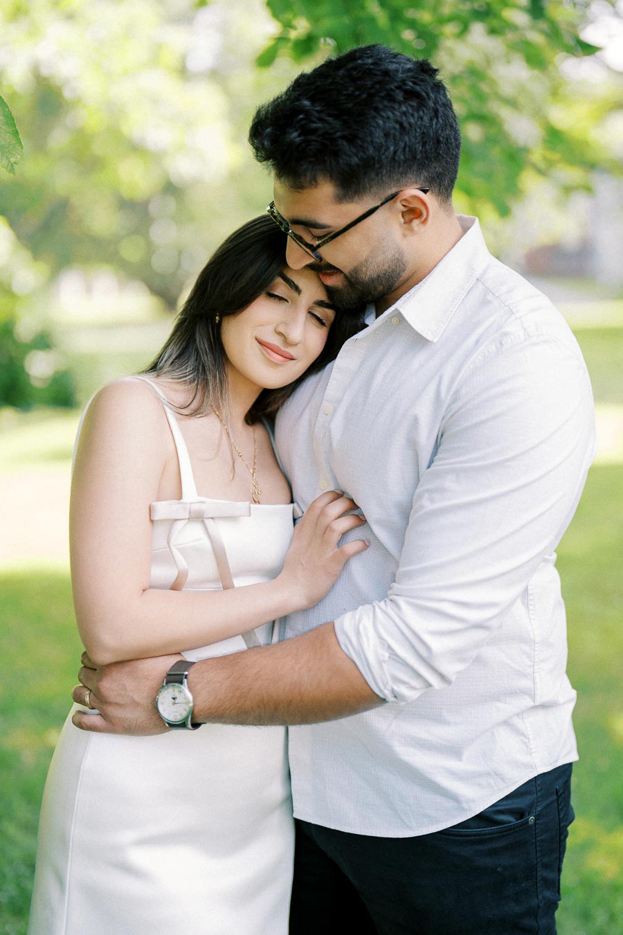 A couple embracing tenderly in a sunny, green park setting, both smiling softly, conveying love and warmth.