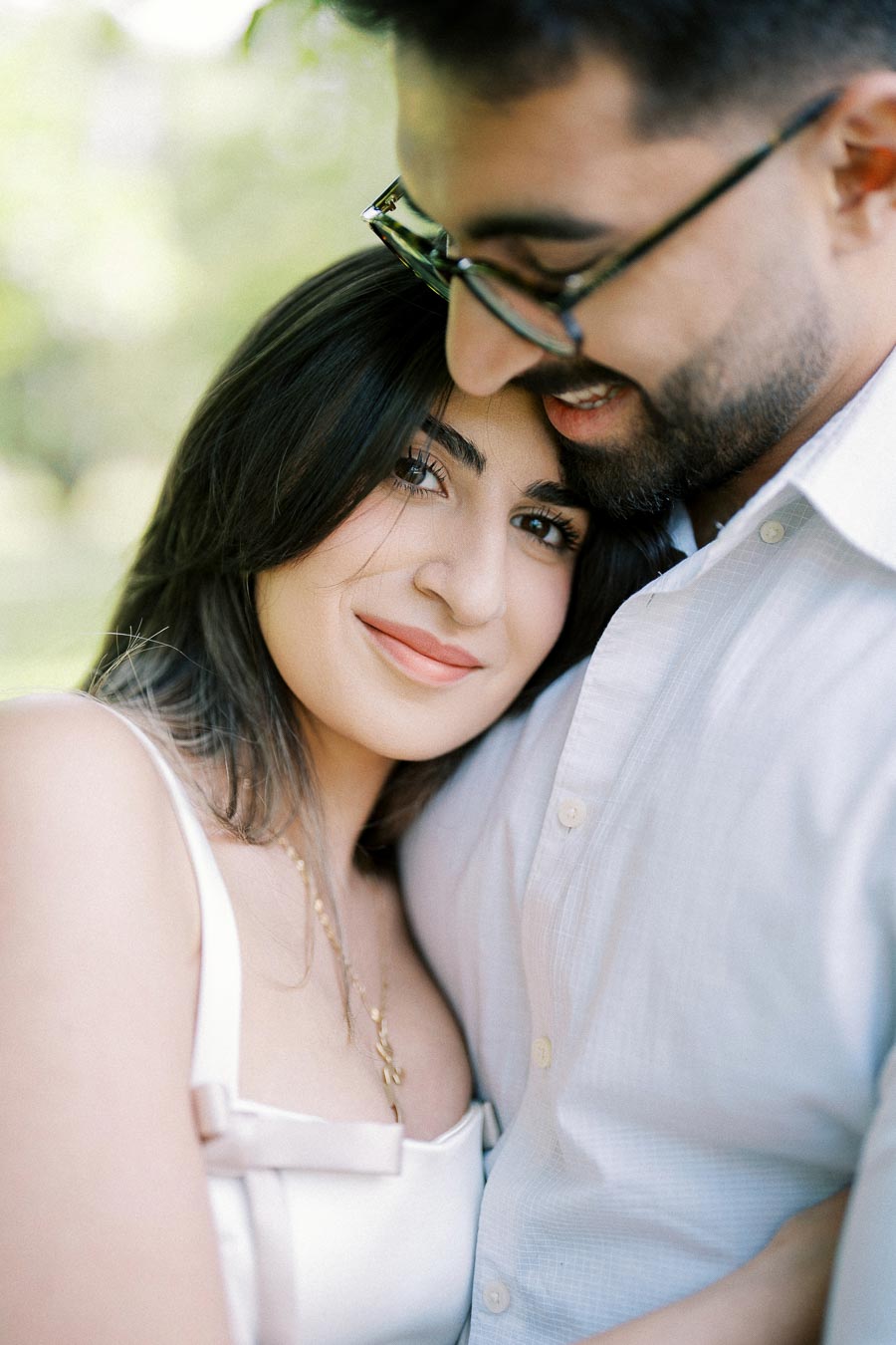 A couple embraces each other in a close, intimate pose, with the woman smiling softly at the camera, outdoors in a lush, green setting.