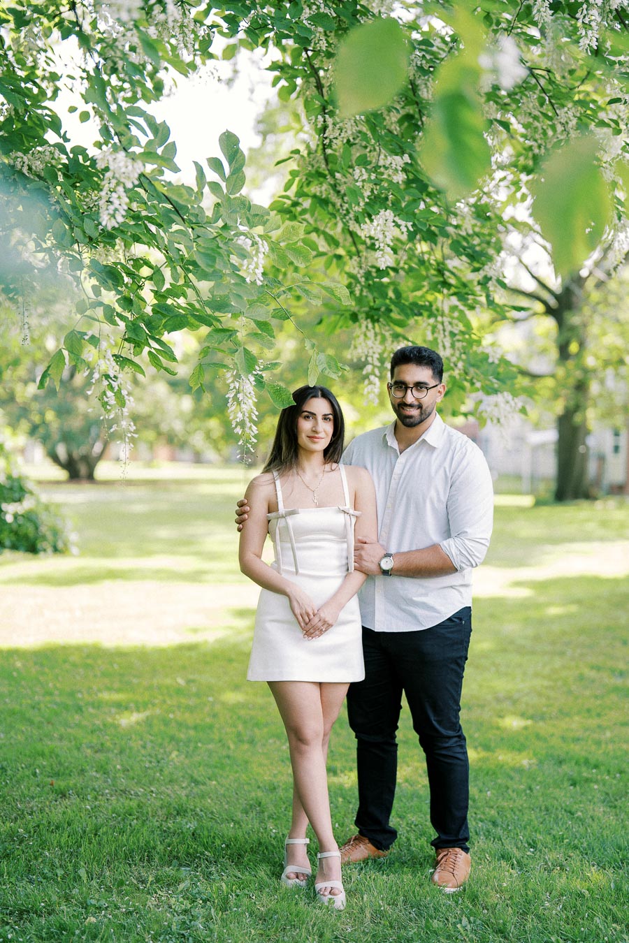 A couple standing together under lush green trees in a park, with the man’s arm around the woman. Both are smiling at the camera, creating a serene and joyful atmosphere. The woman is wearing a white dress, and the man is in a white shirt and dark pants.