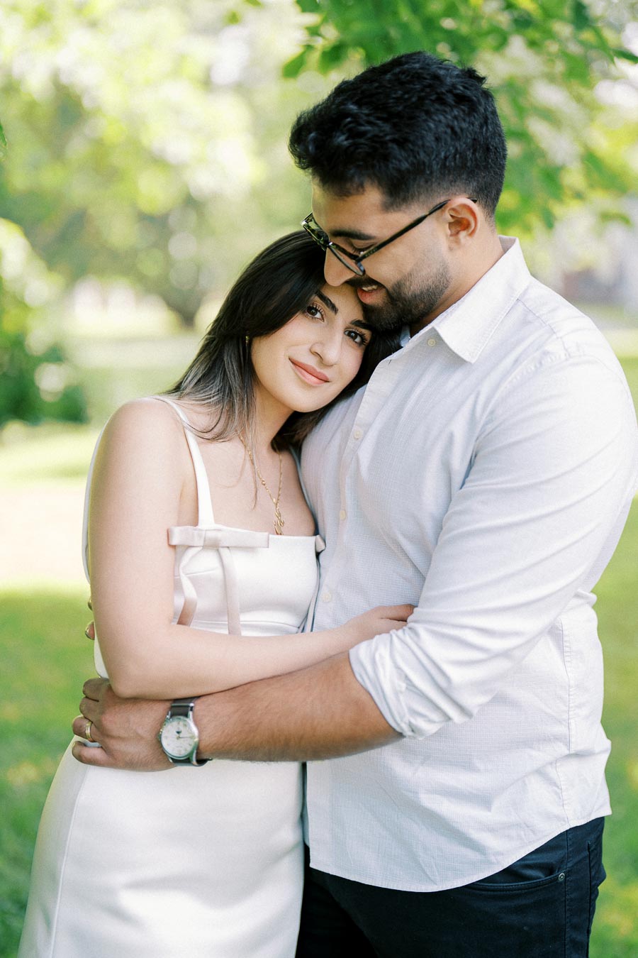 A couple embracing under lush green trees in a sunny park, with the woman leaning her head on the man's shoulder, both smiling and dressed in white attire.